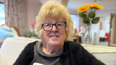 Sue Watson looks at the camera. She has blonde, shoulder-length hair and is wearing glasses. She is wearing a black cardigan over a grey dress. She is sitting on a sofa. A vase with sunflowers is on a table behind her.