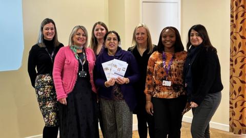 Seven women are smiling whilst they are stood posing for a photograph. The woman in the centre, councillor Jasbir Jaspal, is holding a set of pamphlets.