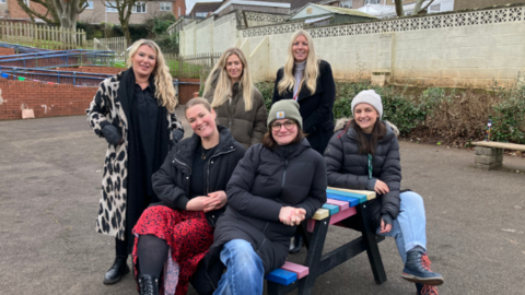 Group of six women sit on and around a picnic bench at Crockerne Primary School in Pill near Bristol, dressed in winter coats with two in beanie hats. They are all smiling. The bench is in the school playground. 