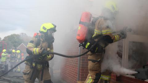 Two firefighters enter a building wearing protective equipment and carrying a hose. A smoke machine is emitting smoke to create a realistic fire environment.
