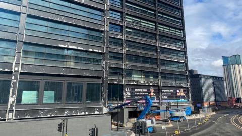 The exterior of a large building with many windows with a grid-like pattern, and grey steel separating the windows. There are lots of white patches on the steel.