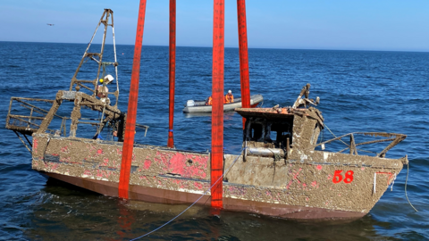 A red boat is being lifted from the sea. There is a small boat in the background.