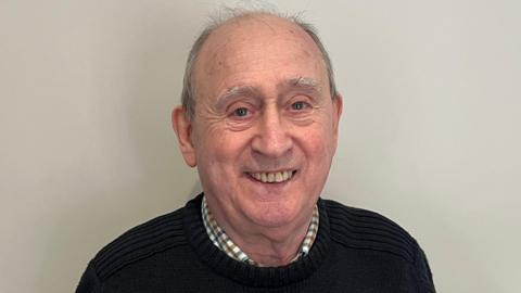 Bill Smith is in the middle of the image looking towards the camera smiling. He has short grey hair and is wearing a black jumper with a shirt underneath. 