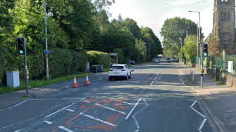 A white car drives along Preston Road near a crossing and two bollards