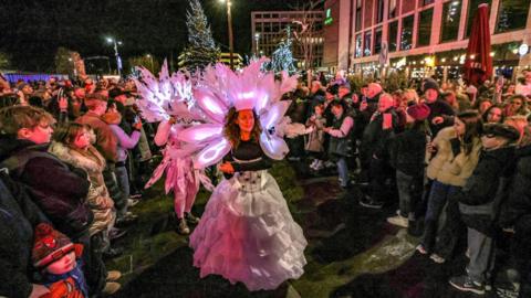 Crowds look on as entertainers make their way to Keel Square as part of the parade. A woman is wearing an illuminated white headdress and flowing white skirt.
