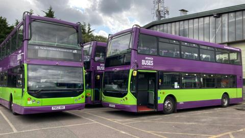 Three purple and lime green buses are parked at a depot. "Ipswich Buses" is displayed on the side of the buses.