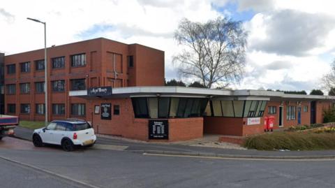 The commercial premises on Well Lane. It is a modern, bricked building with a front entrance. There are cars around. There is a tree in the background and it is overcast.