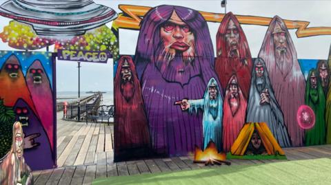 A painting created on Southend Pier showing a number of ghoulish figures, a spaceship and a man hiding in a tent beside his campfire.