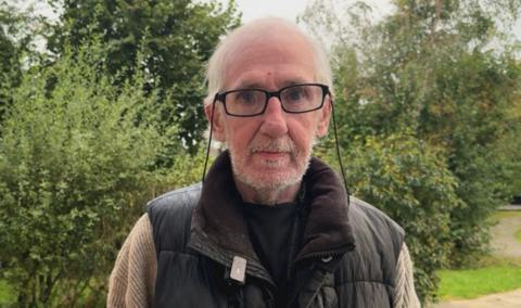 Resident Joe is an elderly man with thinning grey hair and black glasses who has a thin grey beard. He is wearing a dark green body warning over a jumper and hedges are in the background