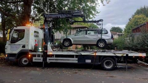 A grey car being lifted onto a white tow truck. Houses and greenery can be seen in the background.