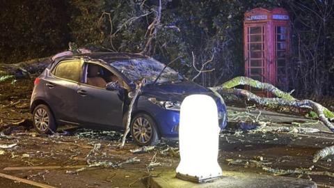 Car with tree branches on it and  smashed windscreen in a road with a phone box.