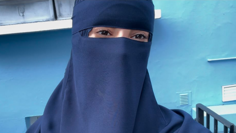 Salsabil in a blue niqab and headscarf, standing in front of a blue wall.