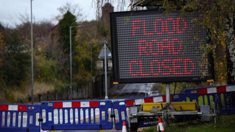 A stock image of a matrix sign warning of a road closed due to flooding. It is placed next to a road that is cordoned off using blue barriers.