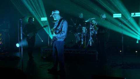 The Snuts perform live on stage. Jack the vocalist/guitarist is clearly visible as he stands in front wearing a light cap, a light jacket and jeans. The other band members are standing behind and aren't so clearly visible. The lights on stage are turquoise.