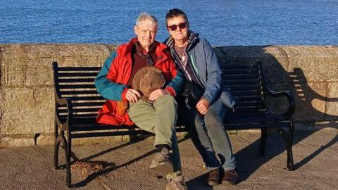 Ruth and her husband sit on a bench by a low stone wall, against a backdrop of the sea. They both wear waterproof coats. Ruth has short dark hair and wears sunglasses.