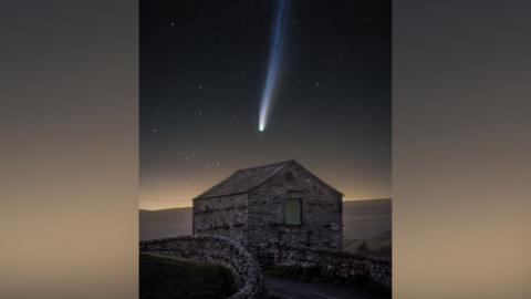 A comet seen in the night sky above a stone house in the Yorkshire Dales. The house is small with just one single window, and is at the side of a road lined by dry stone walls. The comet is a ball of white light in the centre of the photo with a white tail fading to blue towards the top of the picture.