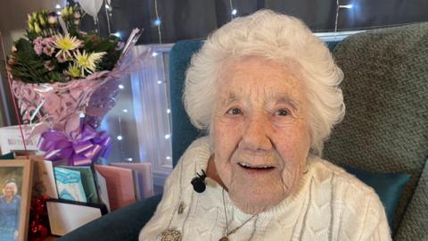 Elsie Wilkins sits in a chair. She has short white hair and is wearing a white cable knit jumper. To her left there are flowers and cards.