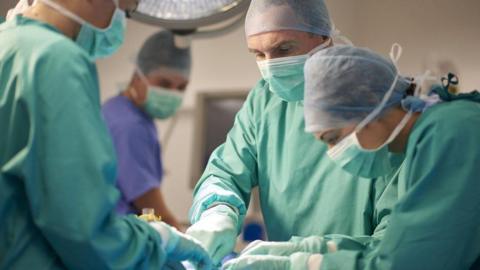 Three surgeons in masks and green gowns operate on a patient who is lying down, unseen