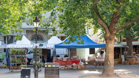 Several market stalls with blue and white pitched roofs are set up on the pavement. They contain tables laden with food and wicker baskets. One has a shelf with potted plants. The weather looks sunny. In the foregound is a tree with green leaves, a black bin and a lamppost