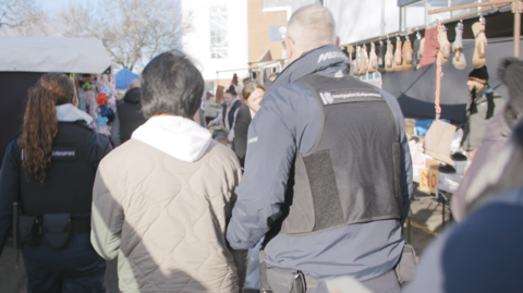 A man in a beige coat and white hooded top is led through a Christmas market by two officers. Only the back of them can be seen. Either side of them are Christmas stalls and shoppers are making their way through the market.