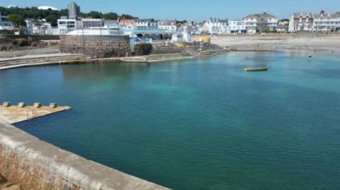 Havre Des Pas bathing pool. The large tidal pool is full or turquoise water. There are steps leading up to an area used for seating and a cafe 