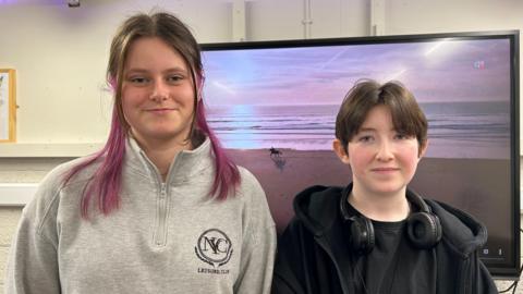 Megan McKenna is on the left, standing next to Cameron Crocker on the right, both looking at the camera with slight smiles. Megan has long brown hair with purple-dyed tips and is wearing a light grey sweatshirt with a zip. Cameron has short brown hair and is wearing a black outfit with a pair of black headphones resting around their neck.