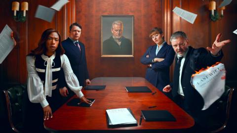 Four people stand around a polished wooden boardroom table beneath a framed portrait, while loose documents swirl through the air and several sheets burn at the edges. Folders and papers labelled “Last Will and Testament” lie on the table, creating a tense, dramatic scene that suggests a dispute.