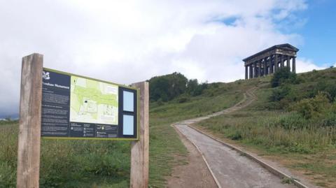 A swerving path leading up to Penshaw Monument, which resembles a Greek temple with stone pillars. There is a National Trust sign at the bottom of the hill with a map and information about the site.