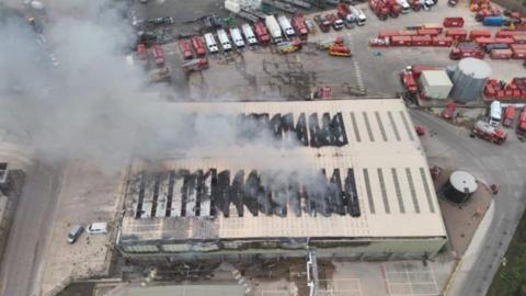An aerial view of the fire at the waste depot. A large rectangular building has got blackened, charred areas and there is smoke billowing from the roof. Lorries are parked around the site with waste management equipment.