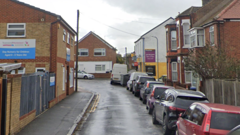 A Google Street picture of a single-track road with terraced housing on one side and a nursery on the other.