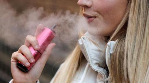 A woman in a white coat uses a pink vaping product and blows out vapour 