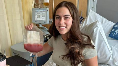 Molly sits in a hospital bed and smiles at the camera while holding a plastic medical bag which contains her stem cells which are a dark pink colour. Behind her is a medical monitor to her left and pillows on her bed to her right. Molly has long dark brown hair with brown eyes.