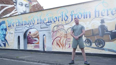 Luke Jenk is wearing shorts and a t-shirt and is standing in front of a mural on a wall in Darlington. It reads We changed the world from here and it celebrates the railway history of the town.