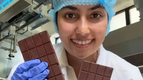 A woman holding two bars of chocolate while smiling into the camera. She is wearing a white gown with purple gloves and a blue hair net.
