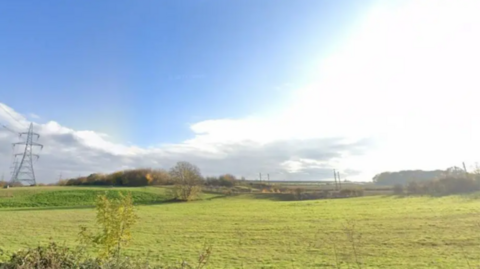 A green field with energy pylons in the left of the image. The field is vast with trees in the background.