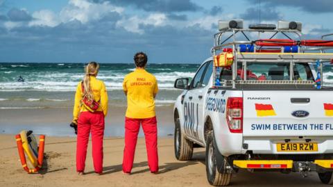 A male and female lifeguard, dressed in red and yellow stood on a beach and looking out to sea next to a RNLI vehicle