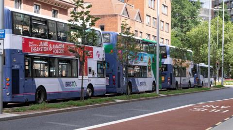 Four First double decker buses lined up on a road in Bristol. There is a bus lane to the right of the image, with large buildings in the background.