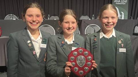 Three young girls in green blazers smiling and Sienna holding a plaque which has silver on it.