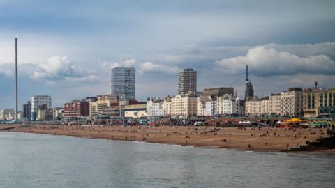 An image of Brighton seafront. A line of Victorian buildings behind a pebble beach with people sitting on it.