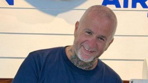 Simon Beston smiling at the camera. He has very short shaved grey hair and beard. He is wearing a blue t-shirt and has a neck tattoo. 