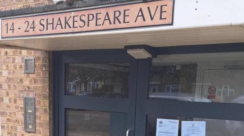 A glass-panelled front door to a block of flats, with a sign above the door reading: "14-24 Shakespeare Ave."