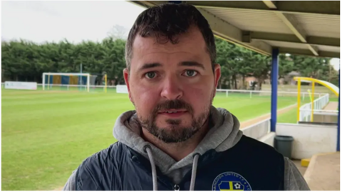 Man with brown hair and brown beard stood in front of a green football pitch looking straight at the camera.