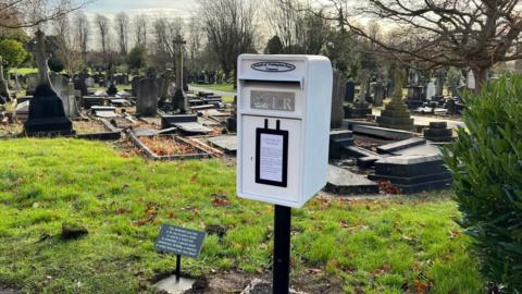 A white post box fitted to a post at Nottingham Road Cemetery. The post box says "Friends of Nottingham Road Cemetery" and "Letters to Heaven". A plaque next to the post box reads: "This dedicated post box is for you to post a letter or card to a loved one to remember a special event, anniversary, birthday, a memory or simply because you miss them."