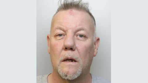 A police mugshot of Ashley Joyner. He is wearing a grey t-shirt and standing against a grey background. He has short grey hair on the sides which is longer and sticking up on the top. He has blue-grey eyes and is looking at the camera with his mouth slightly open. 