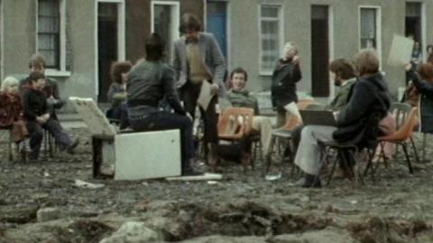 Teenagers sit together on rubbled ground in mismatched chairs and furniture.