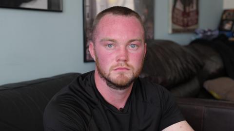 Geraint is facing the camera, wearing a black t-shirt sitting in his living room.