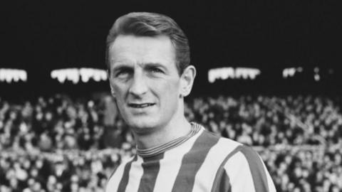 A black and white image of footballer George Eastham, with short dark hair and a striped shirt, during a football match. A crowd can be seen sitting in a stadium in the background.