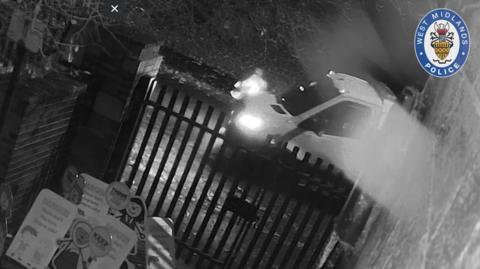 Black-and-white CCTV image of a car colliding with a metal gate at night, headlights on, with debris or dust scattering; West Midlands Police logo visible in the top corner.
