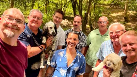 A group of men stand in woods on a sunny evening talking a selfie with two dogs being carried in their arms.