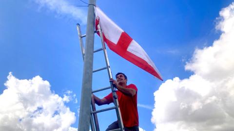 Billy Crotty dressed in jeans and a red t-shirt climbs a ladder to affix a St George's flag to a lamppost